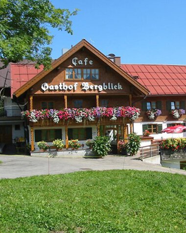 Mountain view inn with flower decorations and terrace in Kleinwalsertal, with a goat in the foreground and hills in the background. | © Kleinwalsertal Tourismus eGen