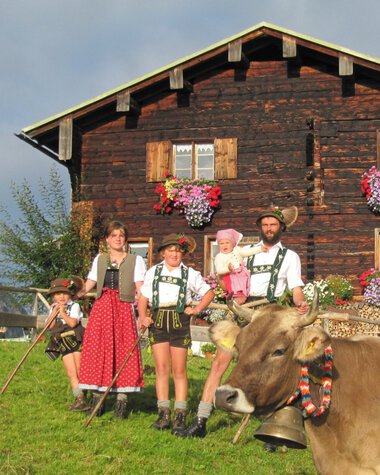 Family in traditional costumes in front of a wooden alpine hut with cows and flowers in Kleinwalsertal, Austria. | © Kleinwalsertal Tourismus eGen