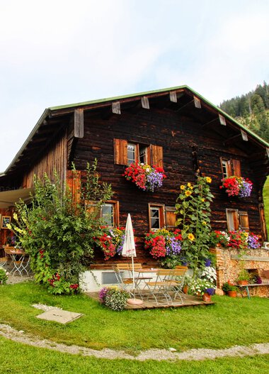 Rustic wooden alpine hut with colorful flower boxes on the windows, surrounded by greenery and with mountain views in the background. | © Ralf Malzer