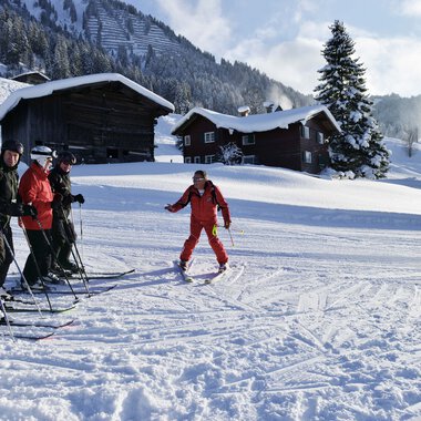 Skilehrer und Teilnehmer beim Skikurs im verschneiten Gebirge, Lehrer zeigt eine Technik, im Hintergrund Hütten und verschneite Bäume. | © Andy Herr