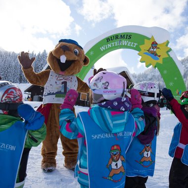 Children in a ski class in Riezlern greeting the Burmi winter world mascot against a snowy mountain background. | © Kleinwalsertal Tourismus eGen • Photographer: Oliver Farys
