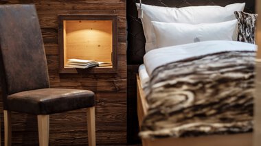 Innenansicht eines gemütlichen Zimmers mit einem Bett, Kopfteil in Schwarz, und einer Holzbox an der Wand, die ein Buch enthält. | © Bergchalet