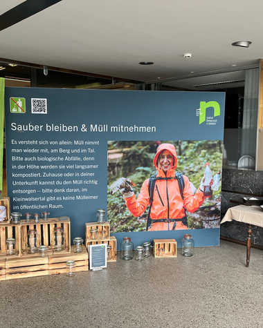 Information booth for the waste campaign featuring a large blue-green poster, wooden boxes, and two tables with tablets indoors. | © Kleinwalsertal Tourismus eGen