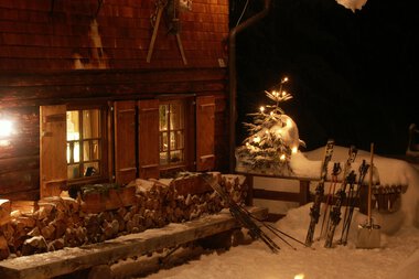 Die Weihnachtshütte bei Nacht mit Schneefall, beleuchtete Fenster und Skiern im Schnee. Weihnachtsdeko mit Lichterkette und Schneemann. | © Kleinwalsertal Tourismus eGen