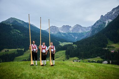 Drei Männer in Trachten stehen in einer grünen Wiese mit Alphörnern, umgeben von Bergen und einer bewaldeten Talregion. | © Kleinwalsertal Tourismus eGen • Fotograf: Oliver Farys
