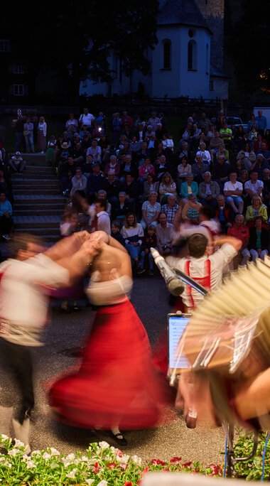 Musiker und Tänzer beim Alphornfestival im Kleinwalsertal, im Hintergrund ein großer Publikum im Freien bei Abenddämmerung. | © Kleinwalsertal Tourismus eGen • Fotograf: Oliver Farys