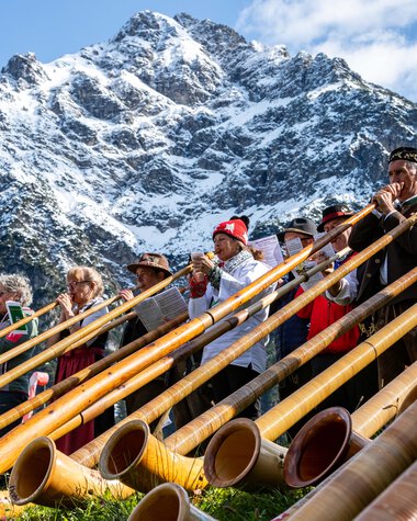 Menschen beim Alphornfestival in den Bergen, mit Berggipfel im Hintergrund, spielen auf traditionellen Alphorninstrumenten. | © Kleinwalsertal Tourismus eGen • Fotograf: Frank Drechsel