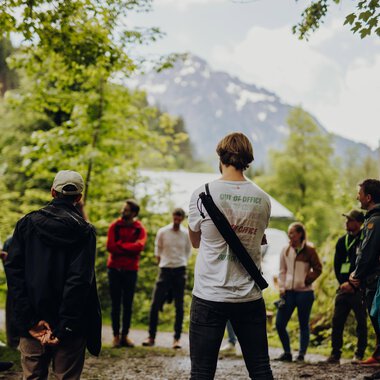 Gruppen von Menschen im Wald, die an einer geführten Tour teilnehmen, mit Berg und Bäume im Hintergrund. | © Kleinwalsertal Tourismus eGen • Fotograf: Philip Herzhoff